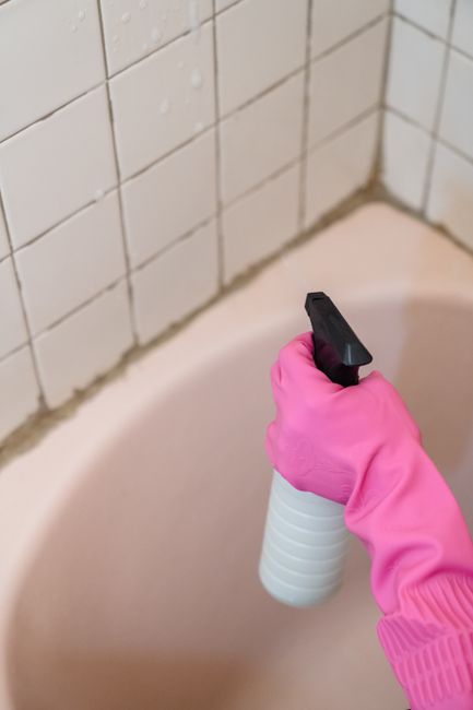 Close-up of a hand wearing a bright pink glove holding a white spray bottle with a black nozzle, positioned over the edge of a porcelain bathtub. The bathtub has a smooth, glossy surface with a gentle